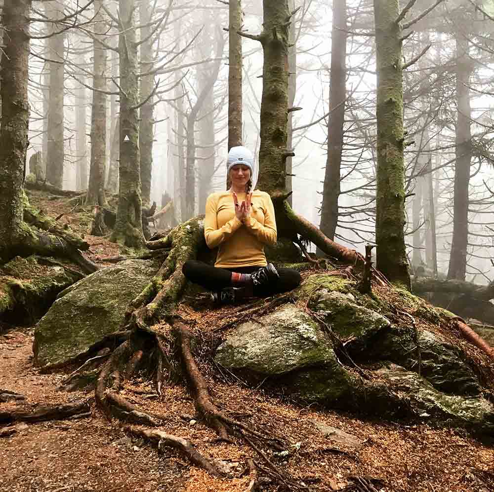 Sara Norman meditating while sitting on a rock ledge in the forest